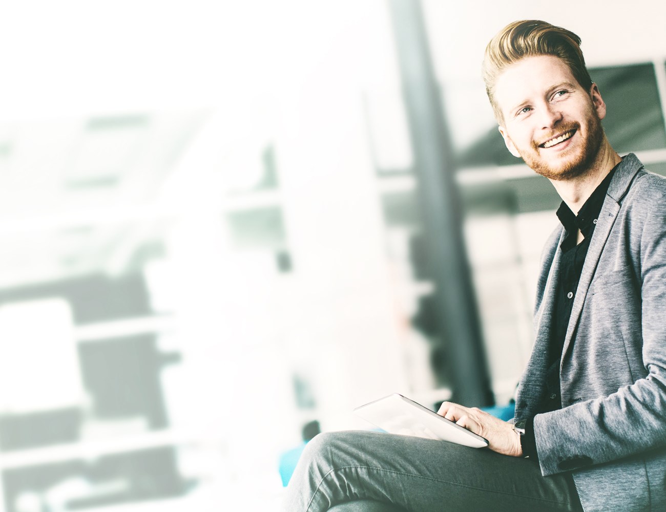 Man in a casual business attire sitting with a tablet and smiling while he works